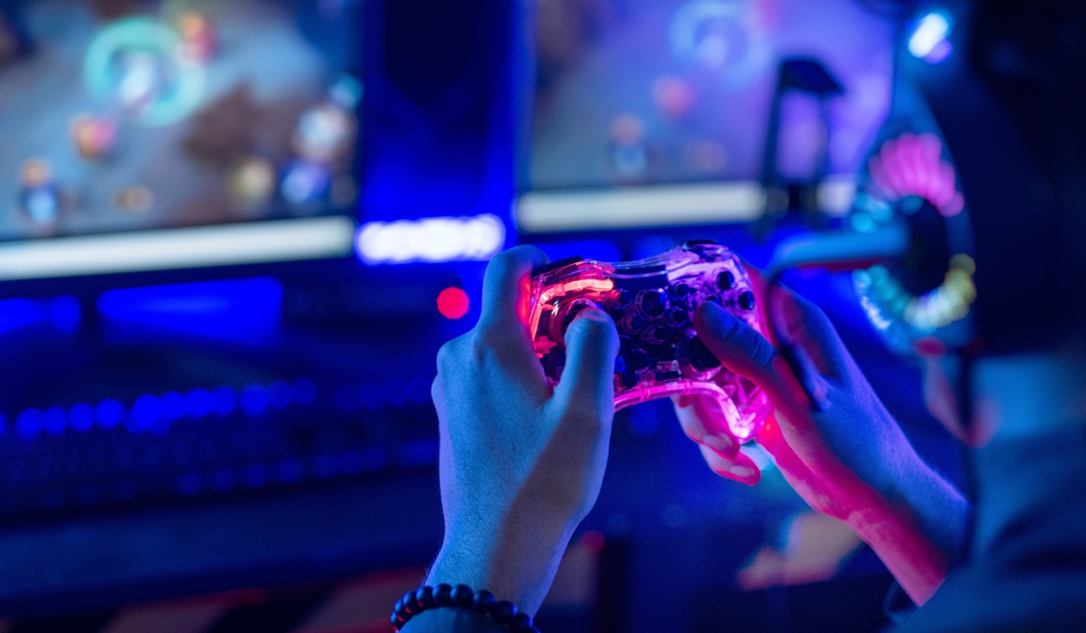 Nahaufnahme-von-Haenden-mit-leuchtendem-Gamecontroller-vor-Monitoren-in-blau-violetter-Gaming-Umgebung Nahaufnahme-von-Haenden-mit-leuchtendem-Gamecontroller-vor-Monitoren-in-blau-violetter-Gaming-Umgebung